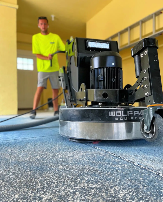 Technician running a Wolf Pack grinder across a blue flake floor in a sunlit space